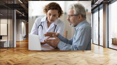 Young female doctor consult elderly male patient in hospital, discuss healthcare insurance on laptop, woman physician talk with mature man client at consultation in private clinic, geriatrics concept Wall mural