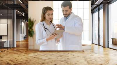 Young female doctor consulting with male colleague, showing patient diagnosis on digital computer tablet in clinic office. Two professional general practitioners using modern tech gadget indoors. Wall mural