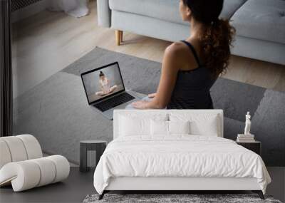 Young female sit on floor at home practice yoga mediate watching video on computer. Woman do stretching exercises take online sports class or training on laptop, have webcam call with instructor. Wall mural