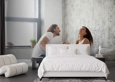 Young happy lovers sitting on bed having fun together, carefree couple laughing spending Sunday morning at home, boyfriend and girlfriend smiling relaxing in bedroom. Good relationship concept Wall mural