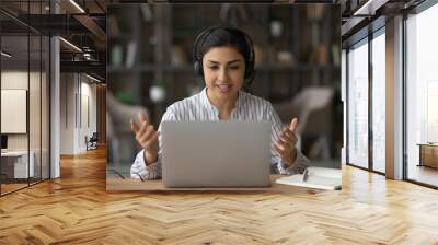 Young Indian woman in headphones sit at desk at home office talk speak on webcam virtual digital event on laptop. Millennial mixed race female in earphones look at computer have video call on gadget. Wall mural