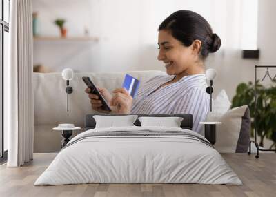 Young indian woman shopper sit on couch enjoy easy paying for goods online using bank card phone. Happy mixed race female traveler book tickets trip hotel at travel agency transport operator website Wall mural