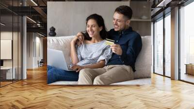 Young millennial Latin couple sit on sofa use laptop holding credit card buying on internet goods for home. Happy online retail web services clients, on-line purchase, instant money transfer concept Wall mural