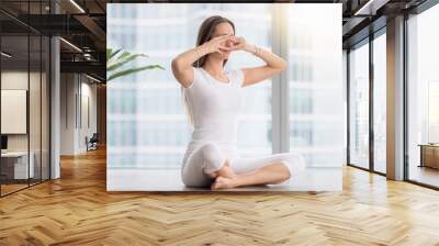 Young woman practicing yoga, sitting in Sukhasana pose, performing Humming Bee Breathing exercise, Bhramari Pranayama technique, working out, wearing sportswear, white t-shirt, pants, full length Wall mural