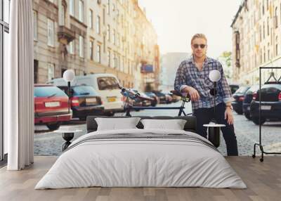 Living in the city, portrait of young man. Young man sitting on the luggage rack of his bicycle in the city while looking into the camera wearing sunglasses Wall mural