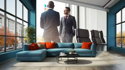 Portrait of two diverse businessmen standing and talking together in ad boardroom, looking over post it note on a window. Wall mural