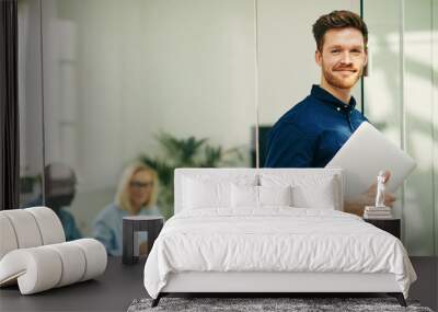 Smiling young businessman standing with his laptop in an office Wall mural