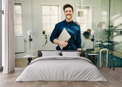 Smiling young businessman standing with his laptop in an office Wall mural