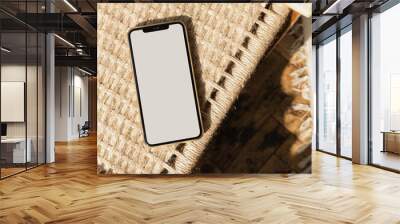 Flatlay of blank screen mobile phone on rattan chair. Flat lay, top view. Copy space mockup template. Warm sunlight shadows. Wall mural