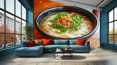 A steaming bowl of Asian noodle soup topped with chopped green onions and a generous amount of spicy chili, served in a black bowl on a wooden table Wall mural