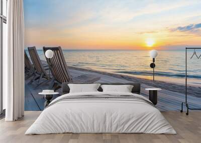 Beach at sunset featuring a row of wooden deck chairs on a boardwalk, overlooking calm ocean waves Wall mural