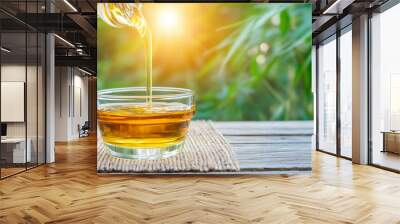 Golden honey being poured into a clear glass bowl on a wooden table, with sunlight and greenery in the background Wall mural