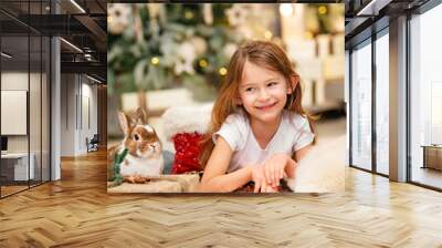 A girl and a rabbit at the Christmas tree with gifts. Christmas mood. A cute little girl smiles at her bunny. Wall mural