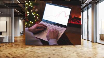 Man freelancer in santa claus hat working on laptop with graphs and charts on screen sitting near christmas tree and fireplace. Wall mural