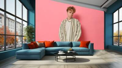 Studio fashion portrait of young confident curly haired ginger man with freckles in trendy clothes green and white striped shirt and chain on neck posing by pink background and looking at camera. Wall mural