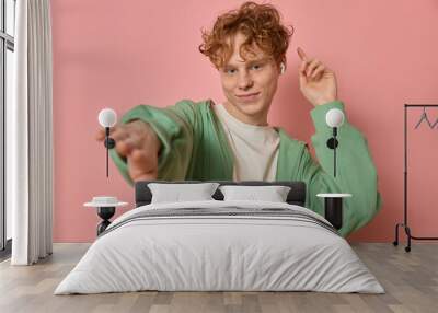 Young caucasian curly haired freckled ginger boy teenager in stylish clothes smiling wearing headphones and enjoying music dancing waving hands over pink background copy space for your advertising. Wall mural