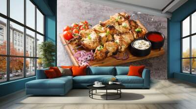 Juicy tasty grilled meat kebabs with fresh vegetables and two sauces close-up on a wooden tray on a gray concrete background. horizontal Wall mural