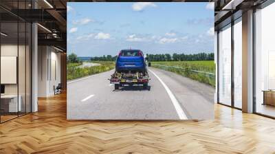 The light truck is a tow truck that carries on itself a blue damaged, broken passenger car on a highway between cities against the background of a blue summer sky with white clouds Wall mural