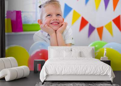 Blonde caucasian boy sits thoughtfully and dreamily at festive table near birthday rainbow cake and makes a wish. Colorful background with balloons. Wall mural