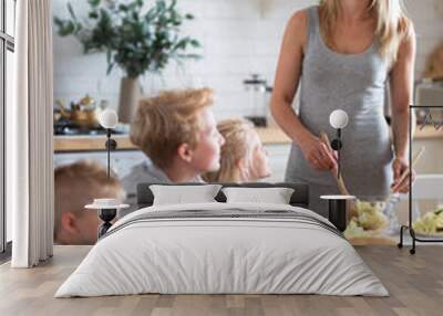 blonde family mother with three kids two sons and daughter eating healthy food in kitchen at home, mom puts green salad on plates to her kids. Wall mural