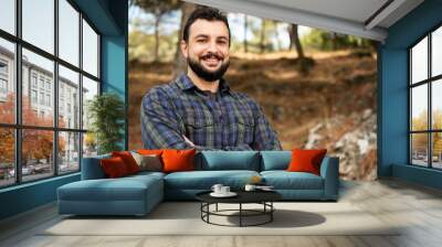 Man with beard and plaid shirt of aesthetic lumberjack crosses his arms in the middle of nature in portrait looking at camera Wall mural