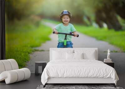 Happy smiling little boy with helmet running and riding on first bike without pedals on sidewalk at city park in warm summer day. Cute 2 years old toddler. Front view. Learning to keep balance. Wall mural