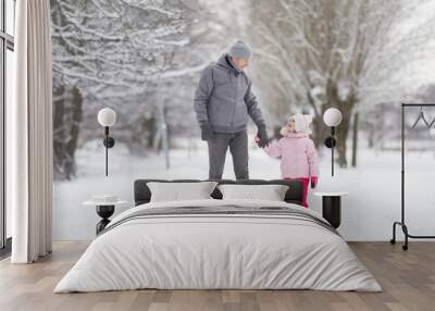 Happy smiling little daughter and young adult father walking on white snow covered sidewalk at park. Spending time together in beautiful cold winter day. Enjoying stroll. Front view. Wall mural