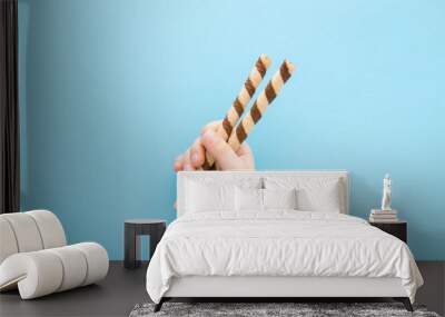 Little child hand holding two striped chocolate wafer sticks on light blue table background. Pastel color. Closeup. Children sweet snack. Top down view. Wall mural