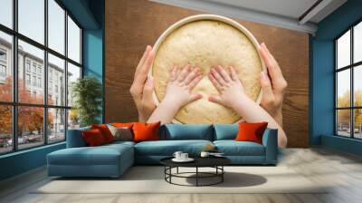 Young adult mother hands holding bowl and baby hands touching fresh white raw dough on brown wooden table background. Spending time together in kitchen. Point of view shot. Closeup. Top down view. Wall mural