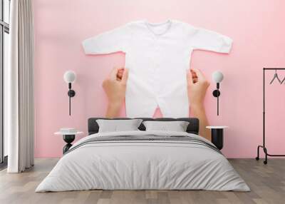 Young adult mother hands holding white new baby pajama with long arms and legs on pink table background. Pastel color. Closeup. Point of view shot. Empty place for text on apparel. Top down view. Wall mural