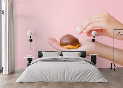 Young adult woman hand holding baked soft bun with chocolate glaze on light pink wall background. Pastel color. Reaching for sweet food. Closeup. Side view. Wall mural