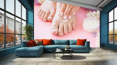 Young adult woman hands applying perfect groomed feet with moisturizing natural cream. Care about clean soft smooth skin. Beautiful branches of colorful lilac. Fresh flowers on pastel pink background. Wall mural