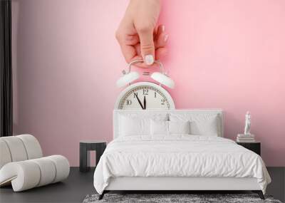 Young woman hand holding white alarm clock on light pink table background. Pastel color. Time concept. Closeup. Wall mural