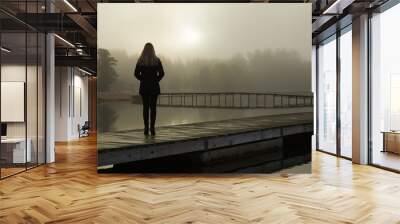 Young woman standing alone on lake footbridge and staring at sunrise in gray, cloudy sky. Mist over water. Foggy air. Early chilly morning. Dark, scary moment and gloomy atmosphere. Back view. Wall mural