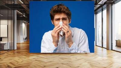 A young man sneezes into a napkin over blue background Wall mural