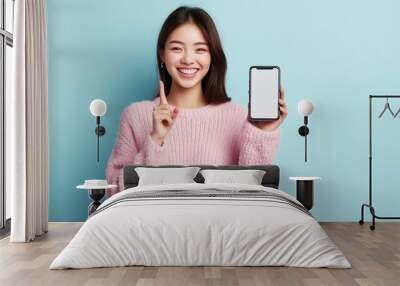 A young woman wearing a cozy sweater smiles brightly while holding up a smartphone against a light blue background Wall mural