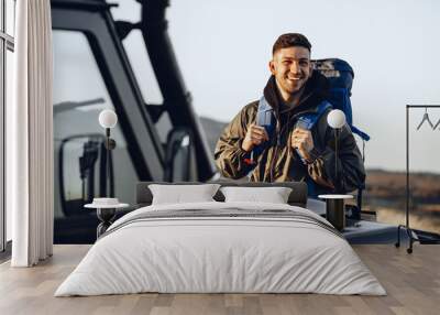 Portrait of a young traveler man in hiking equipment standing near his off-road car Wall mural