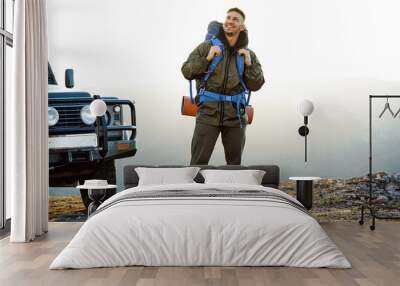 Portrait of a young traveler man in hiking equipment standing near his off-road car Wall mural