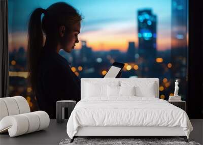 Silhouette of a woman using a tablet against a city skyline at dusk, showcasing vibrant urban lights and a colorful sunset sky Wall mural