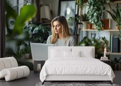 Woman working on a laptop surrounded by greenery in a cozy indoor workspace during daylight Wall mural