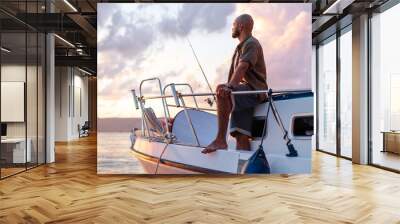 Young african american man standing with fishing rod on a sailboat fishing in open sea on sunset Wall mural