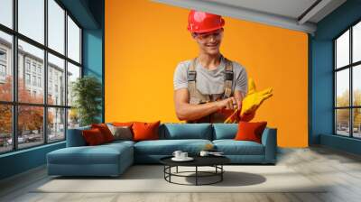 Young construction worker in helmet and uniform posing on yellow background in studio Wall mural