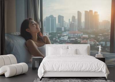 Young woman enjoying sunrise from a hotel room window overlooking a city skyline in the early morning light Wall mural