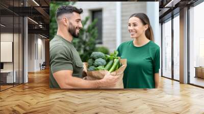 A delivery person handing a bag with fruits and vegetables meal to a smiling woman at the doorstep of her home  Wall mural