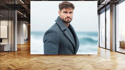 Handsome male model in a gray overcoat standing on a windswept beach waves crashing in the background coat billowing with the wind cool and rugged style  Wall mural