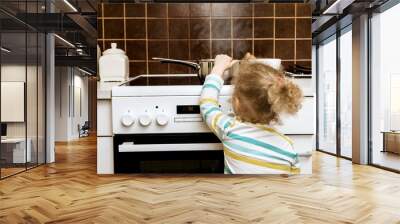 Accident waiting to happen. Toddler girl pulling boiling pot of water over in home kitchen. Hazard at home, skin burn concept( set up, pot is empty). Wall mural