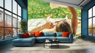 Young woman lying down on a blanket in home yard and sunbathing on a sunny day and reading a real book. Grass and dandilion background, nice vintage look. Wall mural