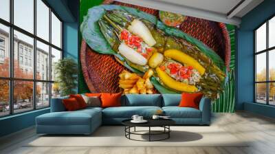 Above view of delicious typical amazonian food, fish cooked in a leaf with yucca and plantain, bowl of salad and fried yucca, served in a wooden plate over a wooden table Wall mural