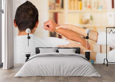 Female physio therapist and man seen from behind, helping patient stretch arm behind head, blurry clinic background Wall mural