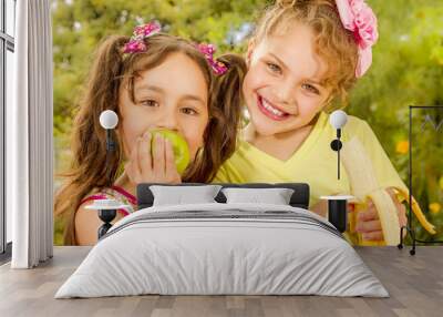 Two beautiful young girls, eating a healthy apple and banana in a garden background Wall mural